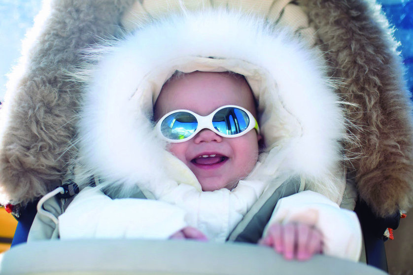 Little baby in warm stroller wearing fur hood snow suit enjoying winter ski vacation in alpine resort. Eyewear and sun protection sunglasses for infants. Child in safe snow goggles in Alps mountains.